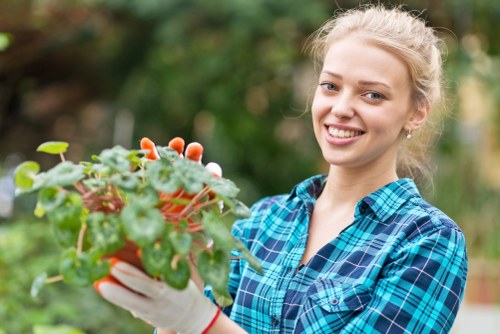 A young woman smiling while holding a potted plant with green and reddish leaves in a garden setting. She is wearing a blue plaid shirt and white gardening gloves, standing in front of a blurred background of trees and shrubbery. The garden features a well-maintained lawn area with dense green grass, bordered by flowering plants and bushes, indicating a healthy outdoor space suitable for gardening services in Victoria. The natural lighting suggests a bright, clear day, highlighting the vibrant foliage and creating a welcoming outdoor environment. This scene reflects typical gardening activities such as planting and garden care in a residential landscape, aligning with services offered by Lawn Mowing Victoria in the local area near Victoria postcodes.