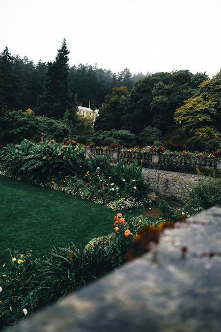 A lush garden scene featuring a well-maintained lawn with neatly trimmed grass bordered by vibrant flower beds containing various colorful blooms and green foliage. In the background, there is a stone