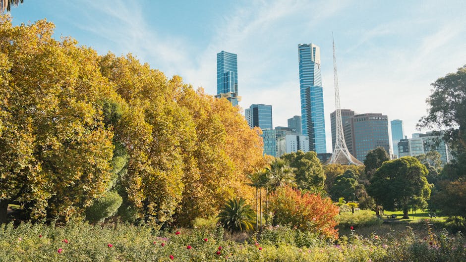A lush, well-maintained garden with a variety of trees and flowering plants in the foreground, including deciduous trees with golden and orange leaves indicating autumn. The garden features neatly tri