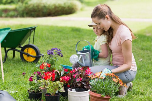 A woman and a young girl are kneeling on a well-maintained green lawn in a backyard garden in Victoria, surrounded by colorful flowering plants in various pots, including purple, red, and orange blooms. They are engaged in gardening activities, with the woman holding a small gardening trowel and the girl wearing green gardening gloves, suggesting planting or tending to the plants. In the background, there is a garden wheelbarrow with a yellow wheel, resting on the grass, and a blurred view of a hedge and trees, indicating a neat and landscaped outdoor space. The scene is lit with natural daylight, and the overall environment appears tidy and lush, showcasing an ideal setting for professional gardening and lawn care services such as those offered by Lawn Mowing Victoria to maintain beautiful, healthy outdoor areas in the local postcode and town.